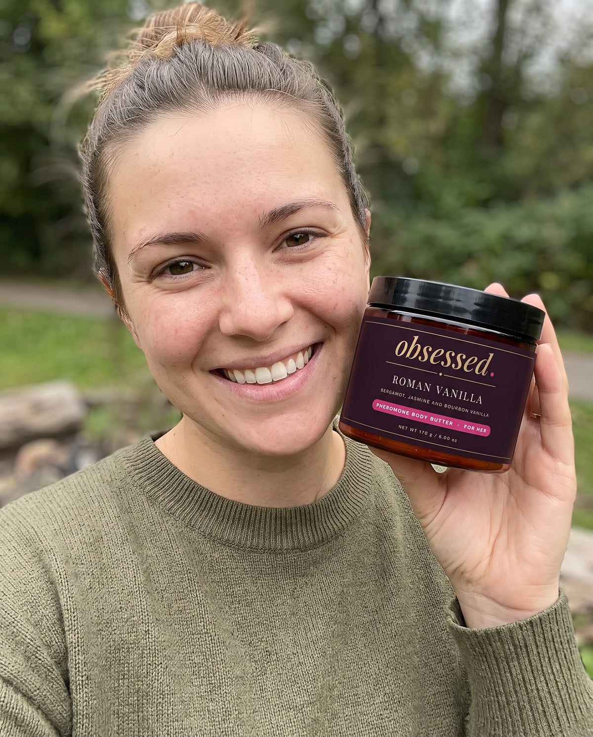 A woman outdoors holds the Roman Vanilla jar beside her face and smiles