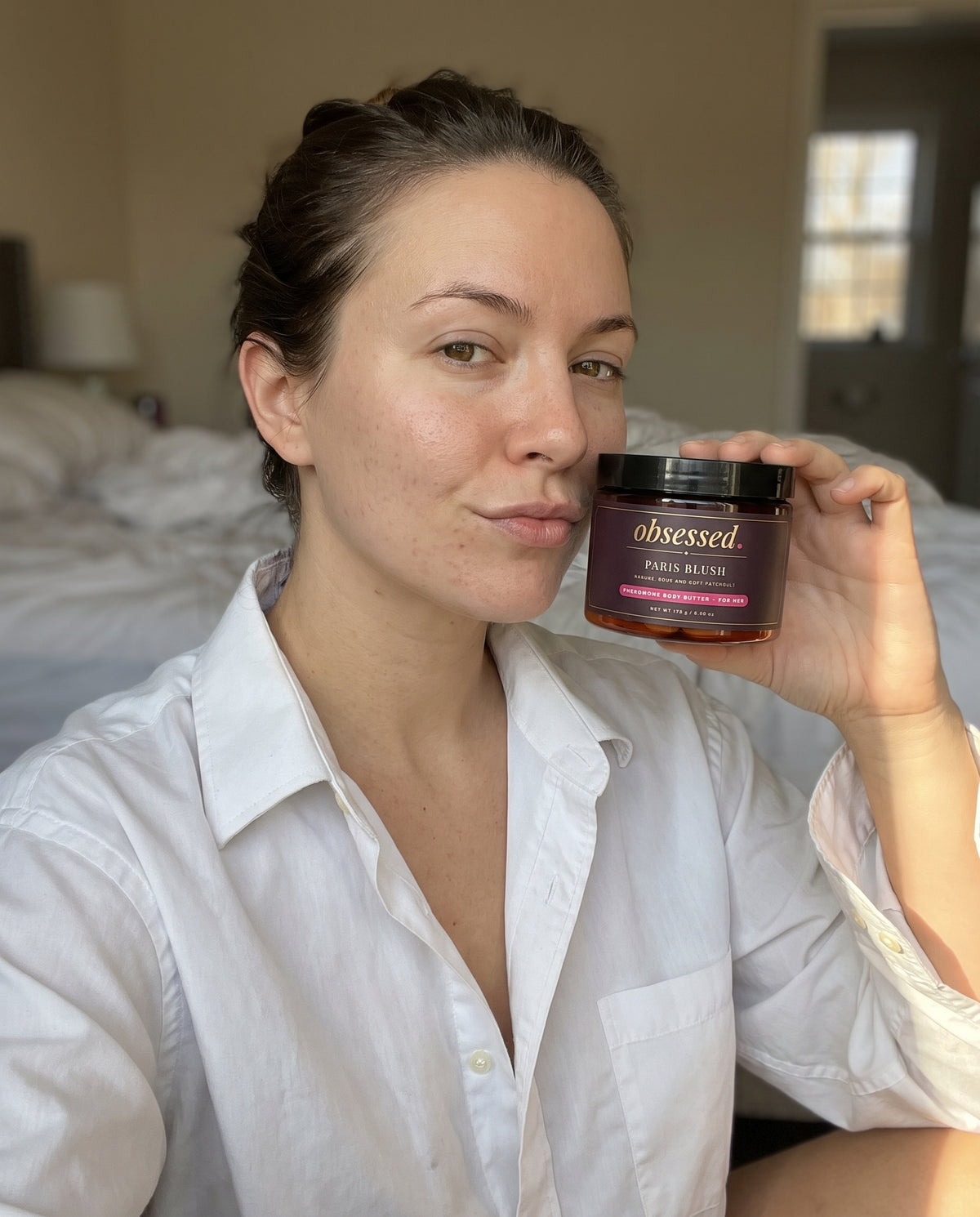 A woman in a white shirt holds the Paris Blush jar beside her face with a smirk