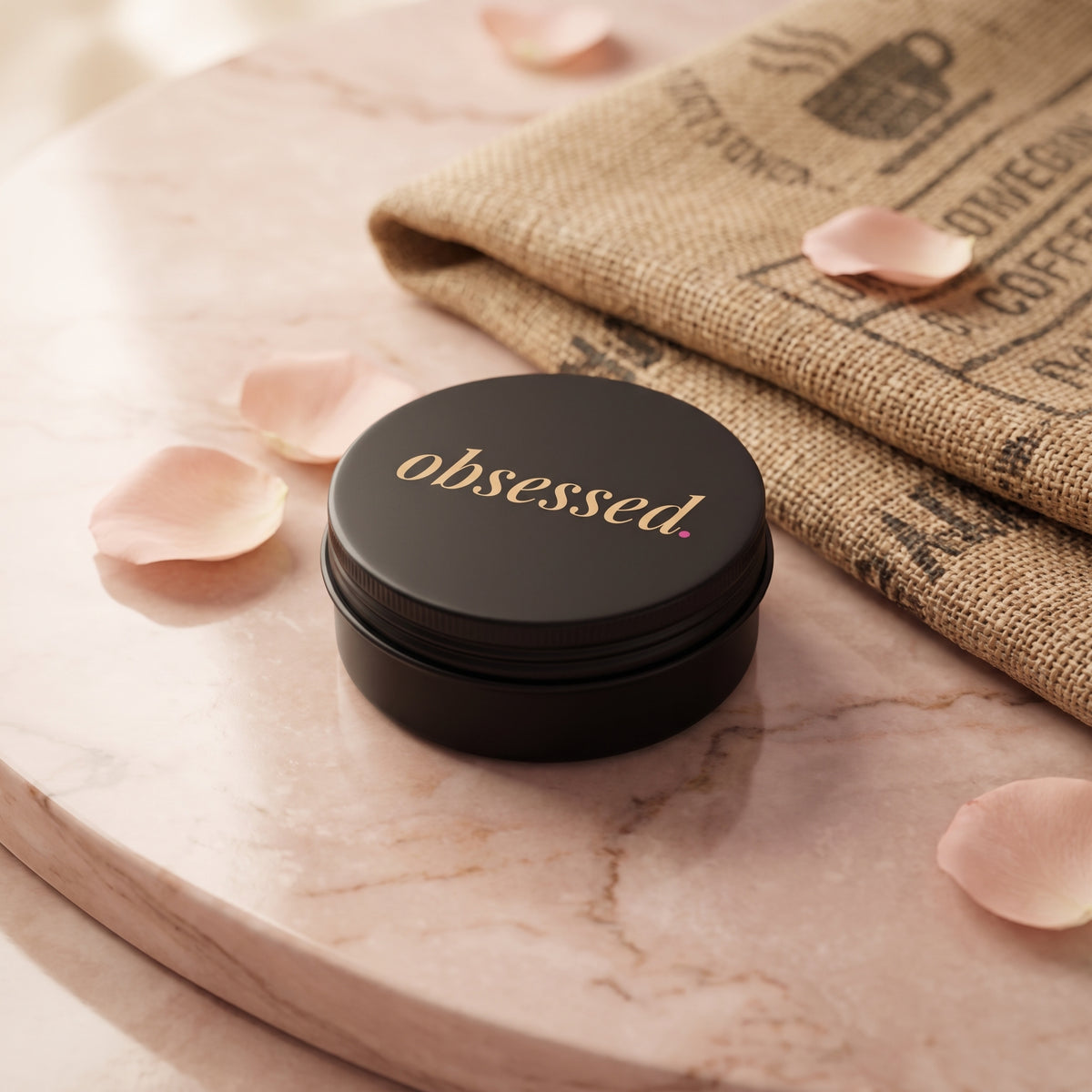 obsessed travel tin: a small matte black tin with the gold obsessed wordmark on the lid, beside rose petals and a coffee sack